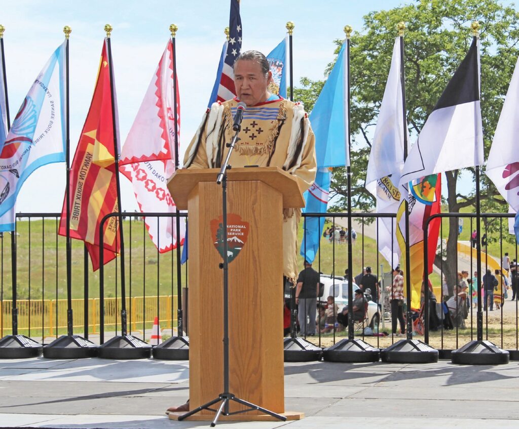 Cheyenne River Sioux Tribal Chairman Ryman LeBeau addresses the crowd at the Little Bighorn commemorative event, speaking from the National Park Service stage as tribal flags stand behind him in a display of respect and presence. (Photo by Ernestine Anunkasan Hopa)